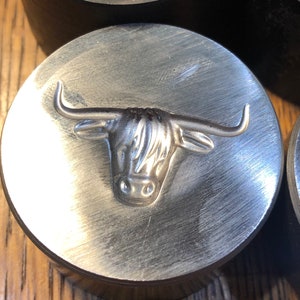 Op de afbeelding: Een zilverkleurige metalen stempel met een gedetailleerd reliëf van een Highland-koeienkop. De stempel is cirkelvormig en heeft een getextureerd oppervlak. De horens en vacht van de koe zijn duidelijk gedefinieerd.