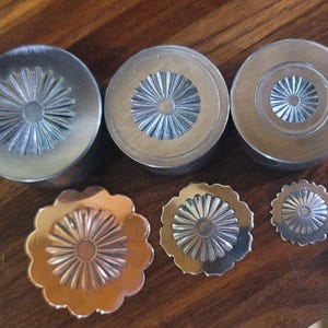 Op de afbeelding: Een verzameling metalen knutselbenodigdheden, waaronder drie cilindrische stukken en drie decoratieve bloemvormige stukken. De stukken hebben een zilveren afwerking en een sunburst-ontwerp in het midden. De items zijn gerangschikt op een houten oppervlak.