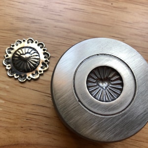 May include: Two metal decorative hardware pieces with a heart and sunburst design. One is a small, scalloped-edged button. The other is a larger, round piece with a brushed metal finish. Both are bronze-toned and sit on a wooden surface.