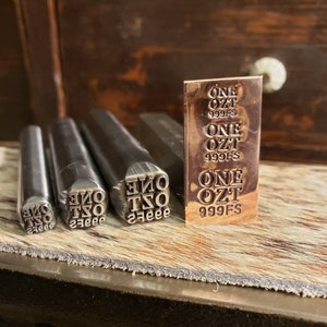 May include: A collection of metal stamps and a small copper bar. The stamps are cylindrical with the text "ONE OZT 999FS" in reverse. The copper bar has the same text stamped on it. The items are on a wooden surface.