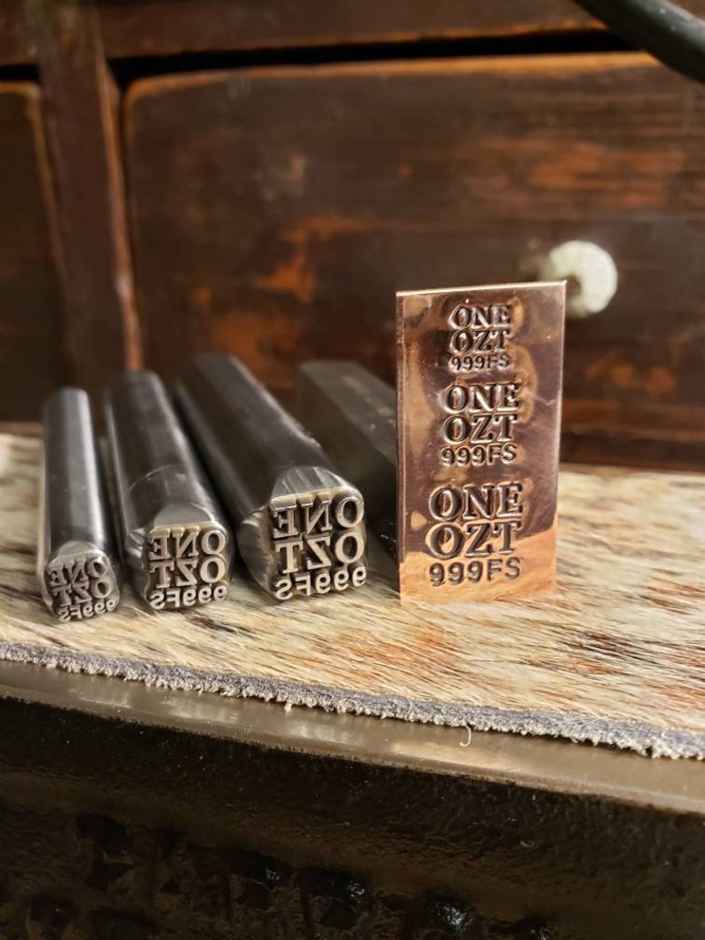 May include: A collection of metal stamps and a small copper bar. The stamps are cylindrical with the text "ONE OZT 999FS" in reverse. The copper bar has the same text stamped on it. The items are on a wooden surface.