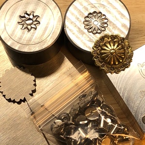 Pode incluir: Close-up de ferramentas de estampagem de metal e ferragens para artesanato em couro. As ferramentas incluem dois selos circulares com desenhos florais, um adorno floral dourado e um saco de rebites de metal. Uma placa de metal com texto também é visível.