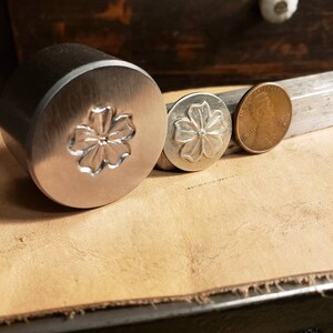 Op de afbeelding: Drie metalen bloemstempels van verschillende formaten worden getoond op een leren oppervlak. De grootste stempel is cilindrisch, de andere zijn cirkelvormig. Een munt ligt naast de kleinste stempel ter vergelijking. De stempels hebben een bloemenontwerp.