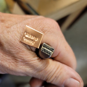 May include: A close-up of a copper stamp and a metal stamp with the text "GRAMS 999ES". The copper stamp is square and the metal stamp is cylindrical. The stamps are held in a hand.