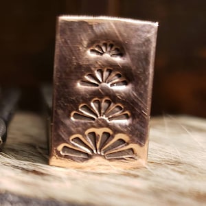 May include: A rectangular copper metal stamp with three fan-shaped designs. The stamp has a textured surface and is set against a blurred background. The fan designs are etched into the metal.