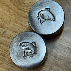 Puede incluir: Dos sellos metálicos circulares con un diseño de pez. Los sellos plateados tienen un relieve detallado de un pez. Los sellos están sobre una superficie de madera.
