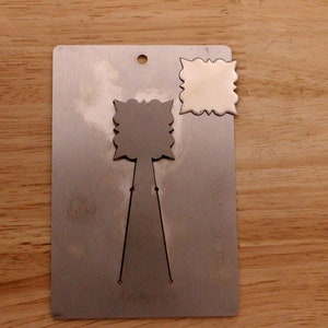 May include: A metal die-cut tool with a decorative design. The tool has a rectangular base with a small hole at the top and a cut-out design. A separate, matching die-cut piece is also present.