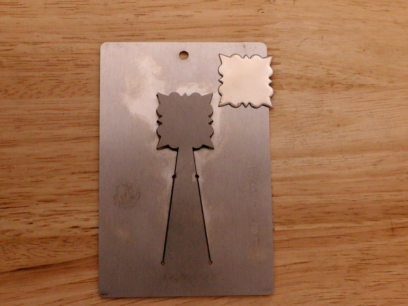 May include: A metal die-cut tool with a decorative design. The tool has a rectangular base with a small hole at the top and a cut-out design. A separate, matching die-cut piece is also present.