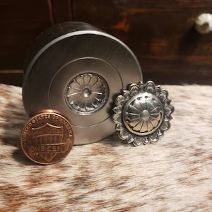 Op de afbeelding: Een zilverkleurig metalen object met een decoratief bloemmotief, naast een kleine, ronde, zilverkleurige knop met een vergelijkbaar ontwerp. Een koperen cent is ook aanwezig, als maatreferentie.
