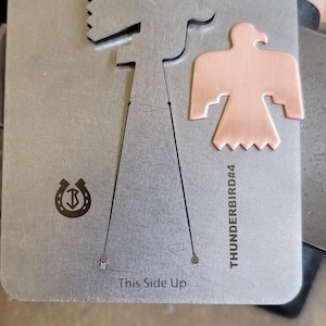 Könnte beinhalten: Metallschablone mit einem Ausschnitt eines stilisierten Vogels und einem separaten kupferfarbenen, vogelförmigen Stück. Die Schablone trägt den Text "This Side Up" und das Kupferstück ist mit "THUNDERBIRD#4" beschriftet. Ein Hufeisen-Logo ist ebenfalls sichtbar.