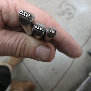 May include: Close-up of four metal stamps with the letters "ase." engraved on the striking surface. The stamps are held in a hand, showcasing the varying sizes of the tools. The stamps are silver in color.