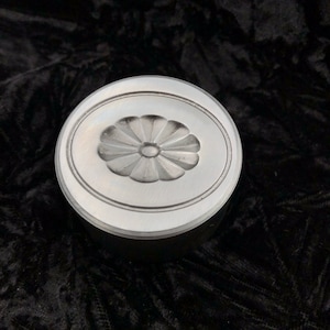 Op de afbeelding: Een zilverkleurige, ronde container met een bloemmotief op het deksel. Het deksel heeft een verhoogde ovale rand en een gedetailleerde bloem in het midden. De container staat op een donkere, getextureerde ondergrond.