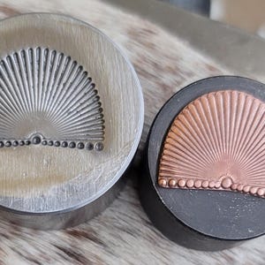 May include: Two metal stamps featuring a fan design. One is silver-coloured, the other copper-coloured. The fan design has radiating lines from a central point, with a row of small circles along the bottom. Ideal for crafting.