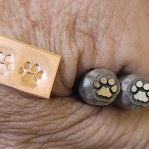 May include: A copper stamp with two paw prints and two metal stamps with paw print designs. The stamps are for creating paw print impressions. The paw prints are a symbol of pets and animals.