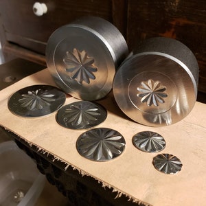 Op de afbeelding: Een verzameling metalen stempelgereedschappen en gestempelde metalen schijven. De gereedschappen zijn cilindrisch met een bloemenontwerp op het platte oppervlak. De gestempelde schijven variëren in grootte en hebben ook het bloemenontwerp. Op een bruin oppervlak.
