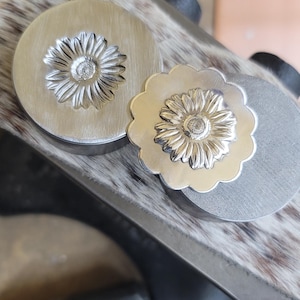 Peut inclure: Gros plan sur deux composants de bijoux en métal argenté en forme de fleur. L'un est rond, l'autre a un bord festonné. Les deux ont un motif de marguerite détaillé au centre. Les composants sont sur une surface texturée.