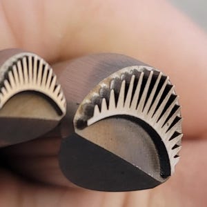 Puede incluir: Primer plano de dos sellos metálicos con un diseño decorativo. Los sellos tienen forma semicircular con un patrón en forma de abanico de líneas verticales y una base curva. El metal tiene un acabado oscuro y mate.