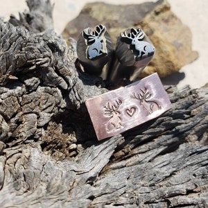 Puede incluir: Dos herramientas de estampado de metal con diseños de ciervos y un rectángulo de cobre estampado con diseños de ciervos y corazones. Las herramientas tienen una silueta de ciervo con cuernos. El rectángulo de cobre muestra dos ciervos con un corazón entre ellos. Los artículos están sobre una superficie de madera envejecida.