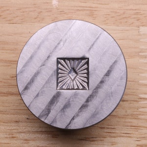 Puede incluir: Objeto met&aacute;lico plateado, circular, con un dise&ntilde;o cuadrado de rayos de sol en el centro. La superficie tiene un patr&oacute;n texturizado y estriado. Probablemente sea una herramienta o un elemento decorativo. Di&aacute;metro: aprox. 5 cm.