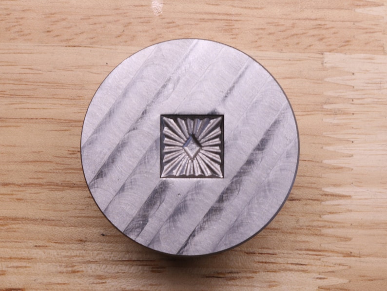 Puede incluir: Objeto met&aacute;lico plateado, circular, con un dise&ntilde;o cuadrado de rayos de sol en el centro. La superficie tiene un patr&oacute;n texturizado y estriado. Probablemente sea una herramienta o un elemento decorativo. Di&aacute;metro: aprox. 5 cm.