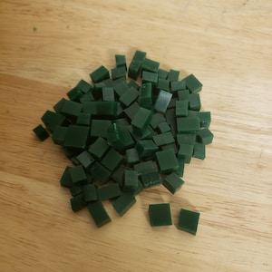 May include: A collection of small, dark green square tiles scattered on a light brown wooden surface. The tiles are uniform in size and shape, suggesting they are intended for a craft or mosaic project. The lighting highlights the smooth texture of the tiles.