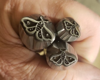 Op de afbeelding: Drie metalen stempels met motontwerpen. De stempels zijn zilverkleurig en voorzien van gedetailleerde motillustraties met steraccenten. De stempels worden in een hand gehouden, wat het ontwerp laat zien.