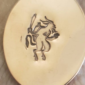 Pode incluir: Pingente oval dourado com uma gravura detalhada de uma criatura mítica. A criatura tem cabeça de cabra, torso humano e segura um bastão. A superfície do pingente é lisa e refletora.