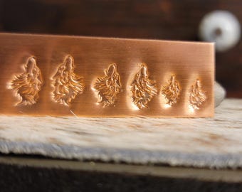 May include: A copper metal stamp featuring a sequence of howling wolf designs. The wolves are finely carved into the metal, showcasing detailed fur and facial features. The stamp is rectangular and has a warm, metallic hue.