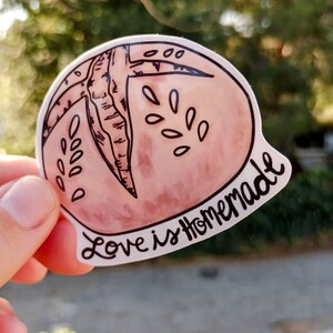 May include: A pink and white sticker with a hand-drawn illustration of a halved grapefruit with seeds. The sticker has the text "Love is Homemade" written in black cursive.