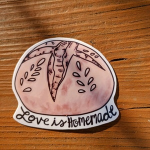 May include: A pink and white sticker with a hand-drawn illustration of a loaf of bread. The text "Love is Homemade" is written in black cursive below the bread.