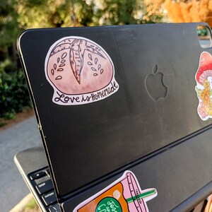 May include: A black laptop with two stickers on the back. One sticker is a pink and brown illustration of a bread roll with the text "Love is Homemade". The other sticker is a pink and orange illustration of a coffee cup with the text "Jesus and Coffee".