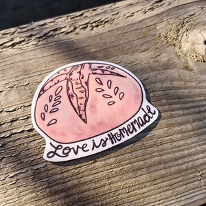 May include: A pink and white sticker with a hand-drawn illustration of a round bread loaf. The text "Love is Homemade" is written in black cursive.