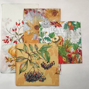 May include: A collection of decorative paper napkins with autumn-themed designs. The napkins feature illustrations of sunflowers, pumpkins, berries, and fall foliage in shades of orange, yellow, green, and red. The napkins are arranged on a white surface.