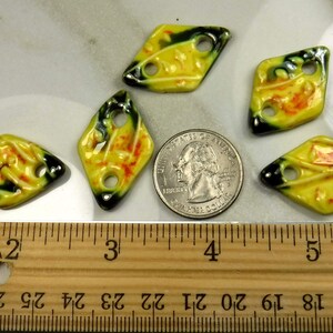 Op de afbeelding: Vijf gele, ruitvormige kralen van keramiek met groene en oranje accenten. De kralen hebben elk twee gaten en zijn ongeveer 2,5 cm in diameter. De kralen zijn gerangschikt op een houten liniaal met inchmarkeringen.