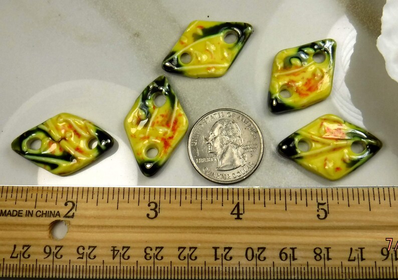 Op de afbeelding: Vijf gele, ruitvormige kralen van keramiek met groene en oranje accenten. De kralen hebben elk twee gaten en zijn ongeveer 2,5 cm in diameter. De kralen zijn gerangschikt op een houten liniaal met inchmarkeringen.