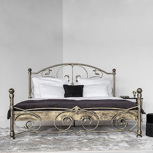 Op de afbeelding: Een gouden metalen bedframe met een sierlijk hoofdeinde en voeteneinde. Het bed is gemaakt van smeedijzer en heeft een decoratief scrollontwerp. Het bed is opgemaakt met witte beddengoed en een zwarte deken.