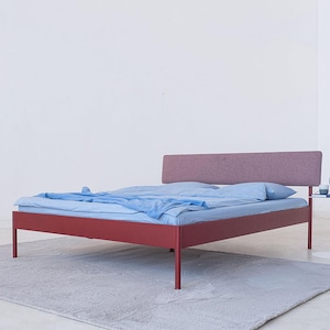 Op de afbeelding: Een modern bedframe met een bordeauxrode metalen basis en een rechthoofdige hoofdbord in een gedempte roze stof. Het bed is opgemaakt met blauwe beddengoed en staat op een grijze vloerkleed. Een kleine bijzettafel met een blauwe lamp staat naast het bed.
