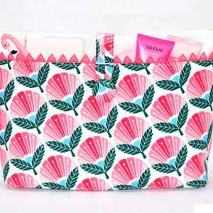 Könnte beinhalten: Eine weiße Canvas-Reißverschlusstasche mit einem rosa-grünen Blumendruck. Die Tasche hat einen rosa Spitzenbesatz und einen schwarzen Riemen.