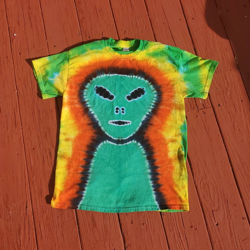Puede incluir: Una camiseta tie-dye con un dise&ntilde;o de cara de extraterrestre verde. La camiseta es una mezcla de naranja, amarillo, verde y negro.