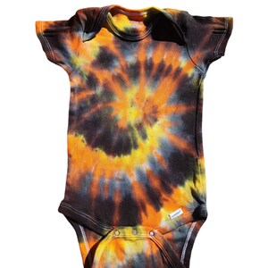 Puede incluir: Un body de bebé de manga corta con estampado tie-dye en negro, naranja y amarillo.