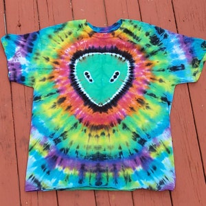 Puede incluir: Una camiseta tie-dye con una cara de extraterrestre verde en el centro. La camiseta es una mezcla de azul, verde, amarillo, naranja, rosa y negro.
