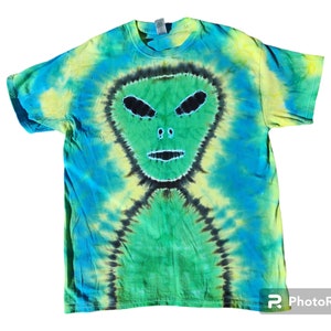 Puede incluir: Una camiseta tie-dye con un dise&ntilde;o de cara de extraterrestre verde. La camiseta es una mezcla de colores azul, verde y amarillo.