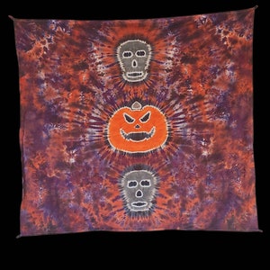 Puede incluir: Una tapicería de pared de tela teñida anudada con una calabaza y dos calaveras en el centro. La tela es una mezcla de naranja, morado y blanco.
