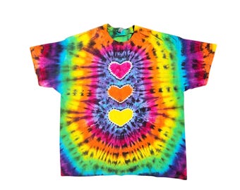 May include: A tie-dye t-shirt with a rainbow swirl pattern and three hearts in a vertical line. The hearts are yellow, orange, and pink.