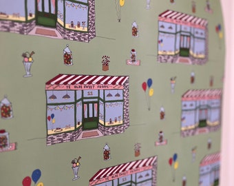 May include: Green wrapping paper with a repeating pattern of a red and white striped awning shop with the text "Ye Olde Sweet Shoppe" and the number "22". The shop has a green door and blue windows. There are also balloons, candy jars, and ice cream cones in the pattern.