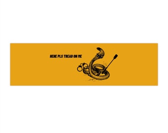 May include: Mustard yellow banner with a black illustration of a coiled snake, handcuffs, and a riding crop. The text "HEHE PLS TREAD ON ME" is printed in black across the top of the banner.