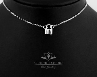 Puede incluir: Un collar de plata con un pequeño colgante de candado. El collar está en un maniquí negro.