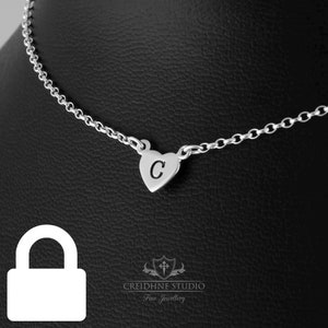 May include: A silver chain necklace with a small heart pendant featuring the letter "C" engraved on it. The necklace is displayed on a black background.