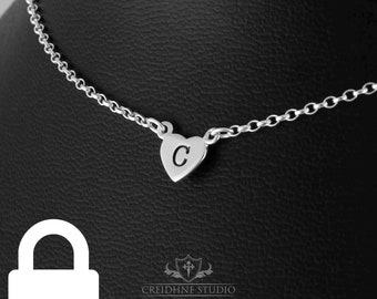 May include: A silver chain necklace with a small heart pendant featuring the letter "C" engraved on it. The necklace is displayed on a black background.