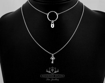 Puede incluir: Un collar de plata con un colgante de llave y un colgante circular con un dije de candado. El collar está en un expositor negro. El texto "CREIDHNE STUDIO Fine Jewellery" está en la parte inferior de la imagen.